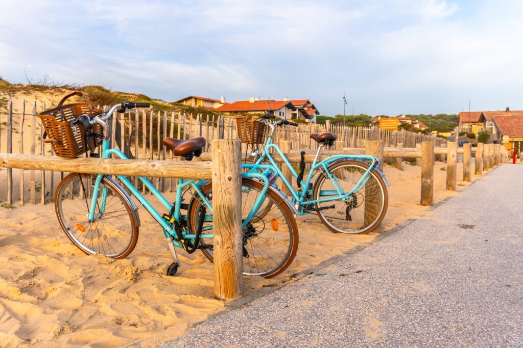 Two vintage style blue bicycles with baskets parked by a wooden fence on sand dunes near a promenade, with holiday homes visible in the background under a soft evening light